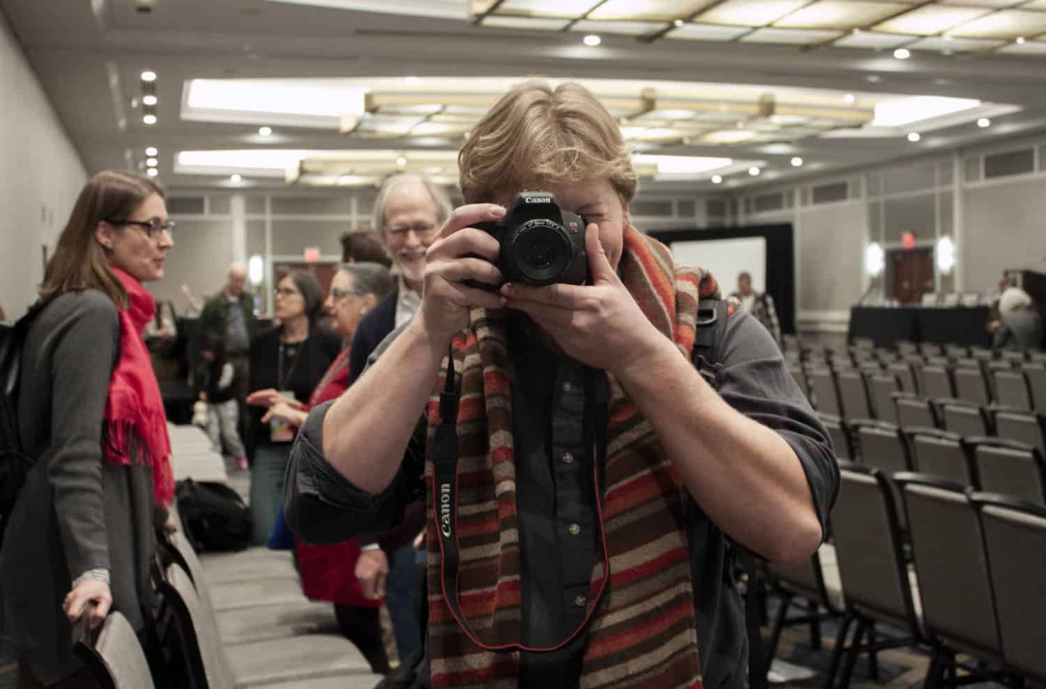 A young man points a camera into the camera in a hotel ballroom. People chat in the background.