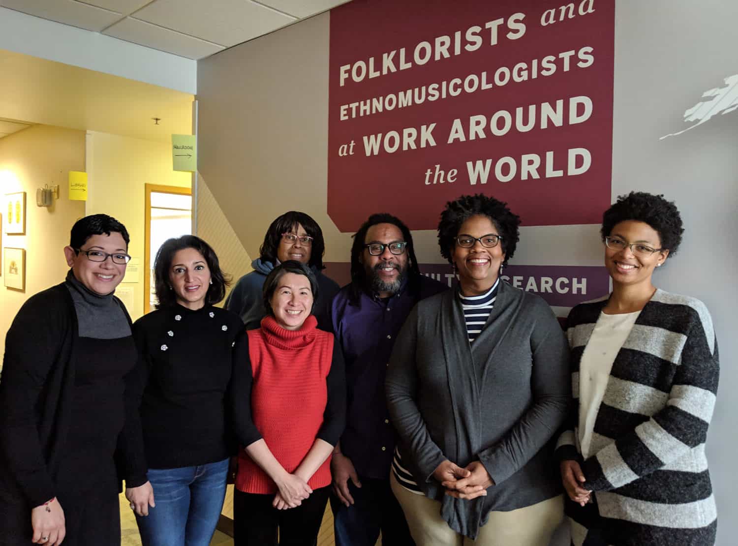 A smiling group stands in front of a sign that reads "Folklorists and Ethnomusicologists at Work Around the World"