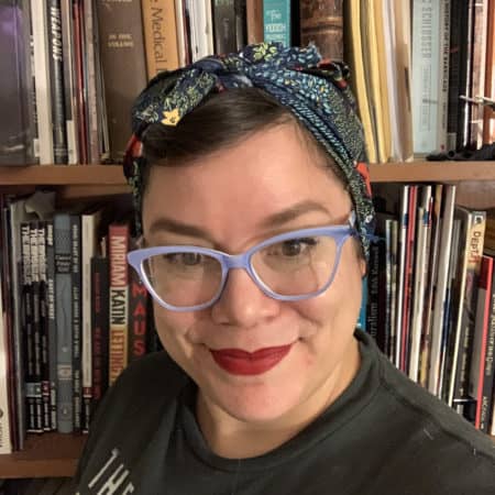A woman, wearing eyeglasses, her hair tied up with a handkerchief, stands in front of a full bookcase, smiling at the camera.