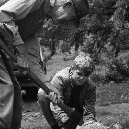 A young boy squats down, watching a elderly man cut away the bark from a log.