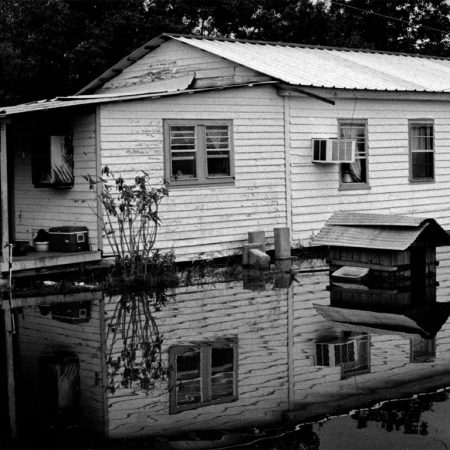 A black and white photo of a small white house standing in water past it's foundation