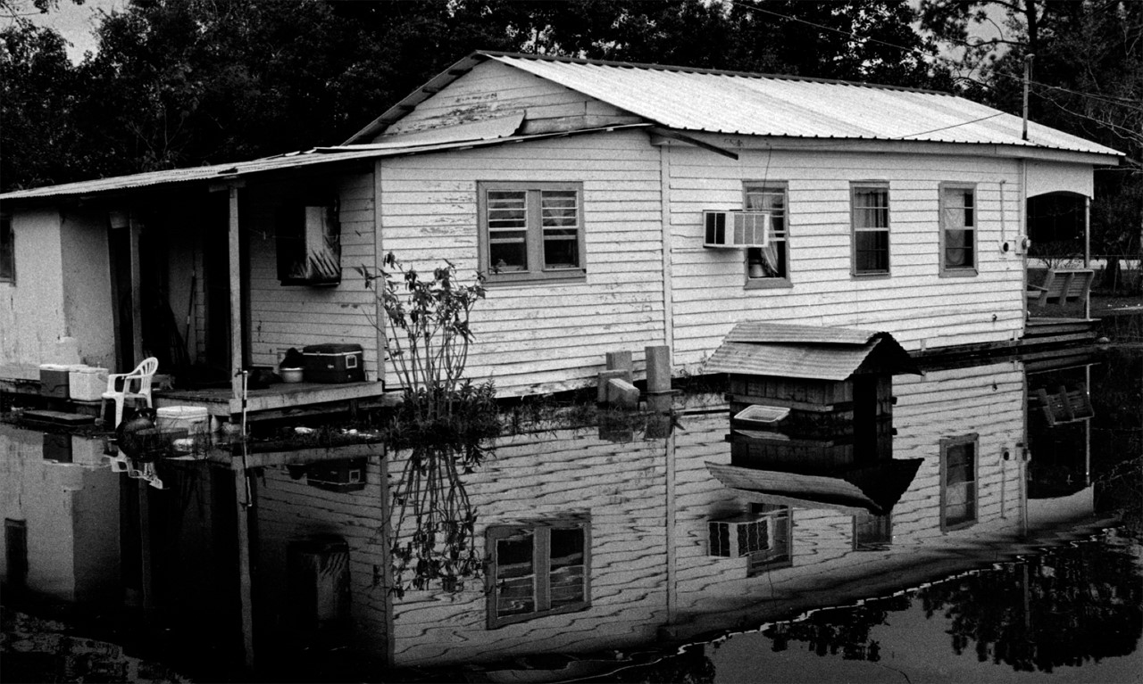 A black and white photo of a small white house standing in water past it's foundation
