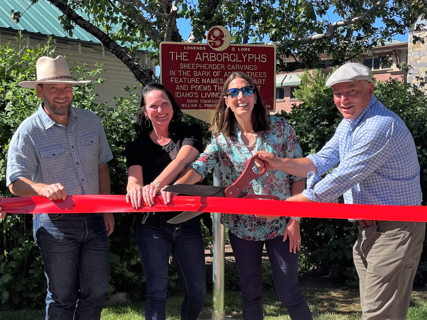 Three people hold a red ribbon while a fourth cuts it with large scissors. They stand in front of a maroon Legends and Lore roadside marker that reads "Legends & Lore / The Arborlyphs (all caps) / Sheepherder carvings in the bark of ... trees feature names ... and poems... Idaho" (some text obscured by people standing in front of the sign)