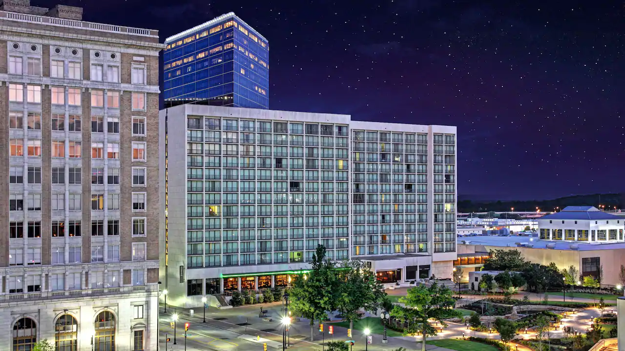 The exterior of the Tulsa Hyatt Regency at night: a modern, building of roughly 12 floors, with a taller high-rise in half of the background behind it, and another to the left. A small park with trees and walkways lies in front of the entrance. The background on the right is a featureless inky blue sky.