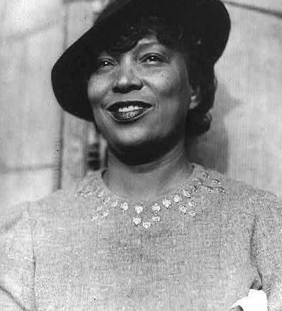 Black and white headshot of African American folklorist, Anthropologist, novelist, and dramatist Zora Neale Hurston. Hurston is wearing a stylish hat that is slanted to the right and a long-sleeved dress with gemstones applied to the collar in a semi-circular pattern. Hurston is smiling slightly, wearing lipstick, and looking up and out of the frame.