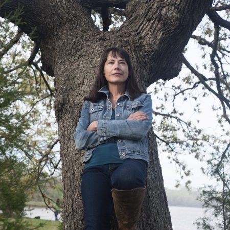 Photo of Mish leaning against a large tree. Mish's left leg is propped against the tree, and is wearing a jean jacket and dark blue jeans.