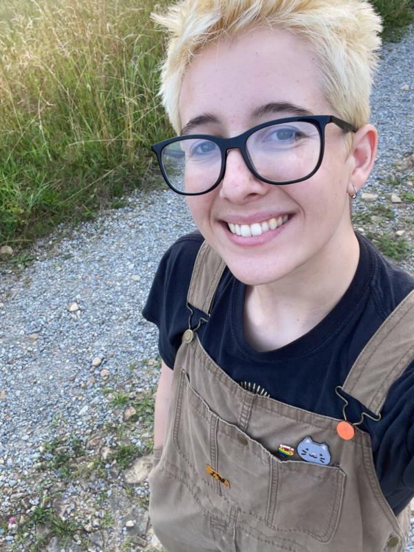 photograph of ahlstone on a dirt road wearing tan overalls, black-rimmed glasses, and smiling.