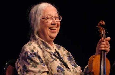 vivian williams, who is wearing a tan shirt with violet flowers and wears glasses, smiling and holding a fiddle.