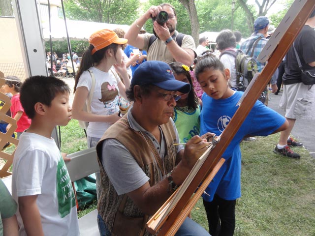 An artist who is a Peruvian man in a blue baseball cap paints at a wooden easel surrounded by four interested children. A man with a camera is capturing the scene. the artist is working intently with a small brush. The children are captivated by his work.