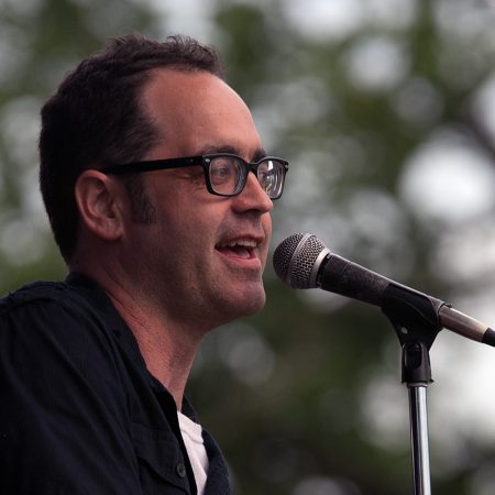 cliff murphy, who wears glasses and a back shirt, singing into a microphone with a tree behind him