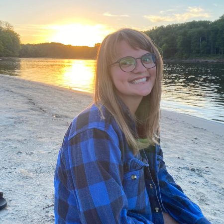 photo of samantha ruth brown sitting on a beach smiling while wearing a blue and black flannel checkered shirt