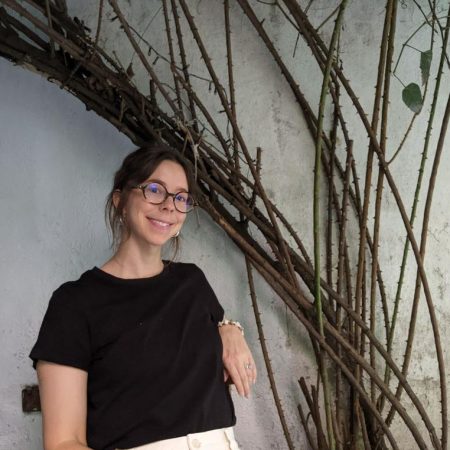 Jordan looks into the camera smiling. She is a white woman with brown hair and round glasses. There are vines crawling up the wall behind her.