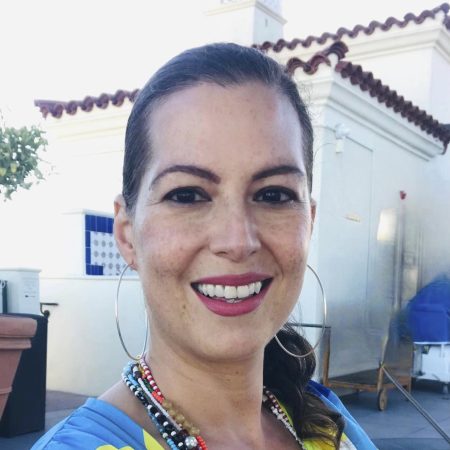 Cuban-American scholar Elizabeth Pérez is pictured here with brown hair pulled back into a ponytail. She is smiling and wearing a rainbow-striped wrap, silver hoop earrings, beaded necklaces, and a pin that says "She/Her."